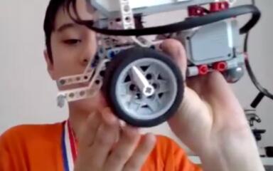 Хлопчик у помаранчевій сорочці з медаллю тримає робота в стилі LEGO, показуючи його колеса та проводку зблизька, з гордістю демонструючи свій проект.